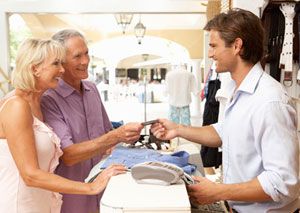 An older couple stands at a store counter, smiling as the man hands a credit card to a young male cashier. Clothes and a credit card terminal are visible on the counter in a bright, open shop.