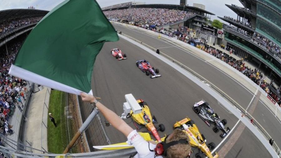 A race official waves a green flag to start an IndyCar race, with cars speeding down the track and a packed grandstand of spectators in the background.
