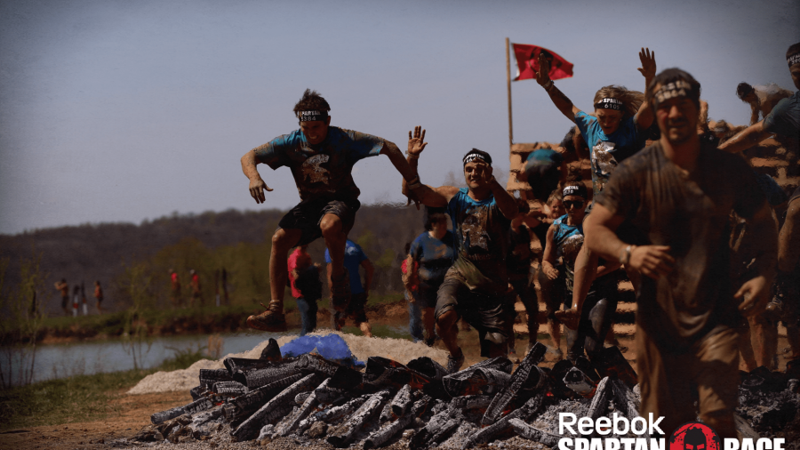Group of people in athletic gear and headbands leap over burning logs during a muddy outdoor obstacle course race, with a Reebok Spartan Race logo in the bottom right corner.