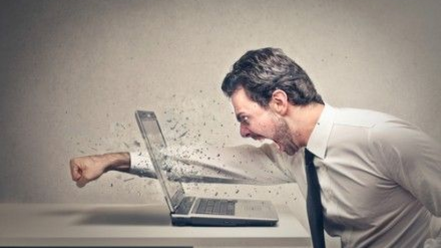 A man in a dress shirt and tie angrily punches a laptop, causing the screen to shatter and fragments to fly, while sitting at a desk against a plain background.
