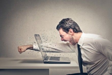 A man in a dress shirt and tie angrily punches a laptop, causing the screen to shatter and fragments to fly, while sitting at a desk against a plain background.