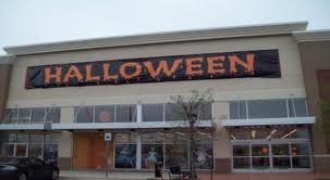 A large store with a black banner above the entrance that reads HALLOWEEN in orange, festive letters. The storefront has glass windows and doors, with some Halloween decorations visible inside.