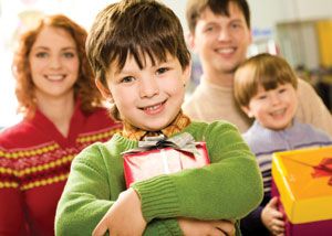 A smiling young boy in a green sweater holds a wrapped gift, standing in front of a smiling woman, man, and another young boy who is also holding a present. The group appears happy and festive.