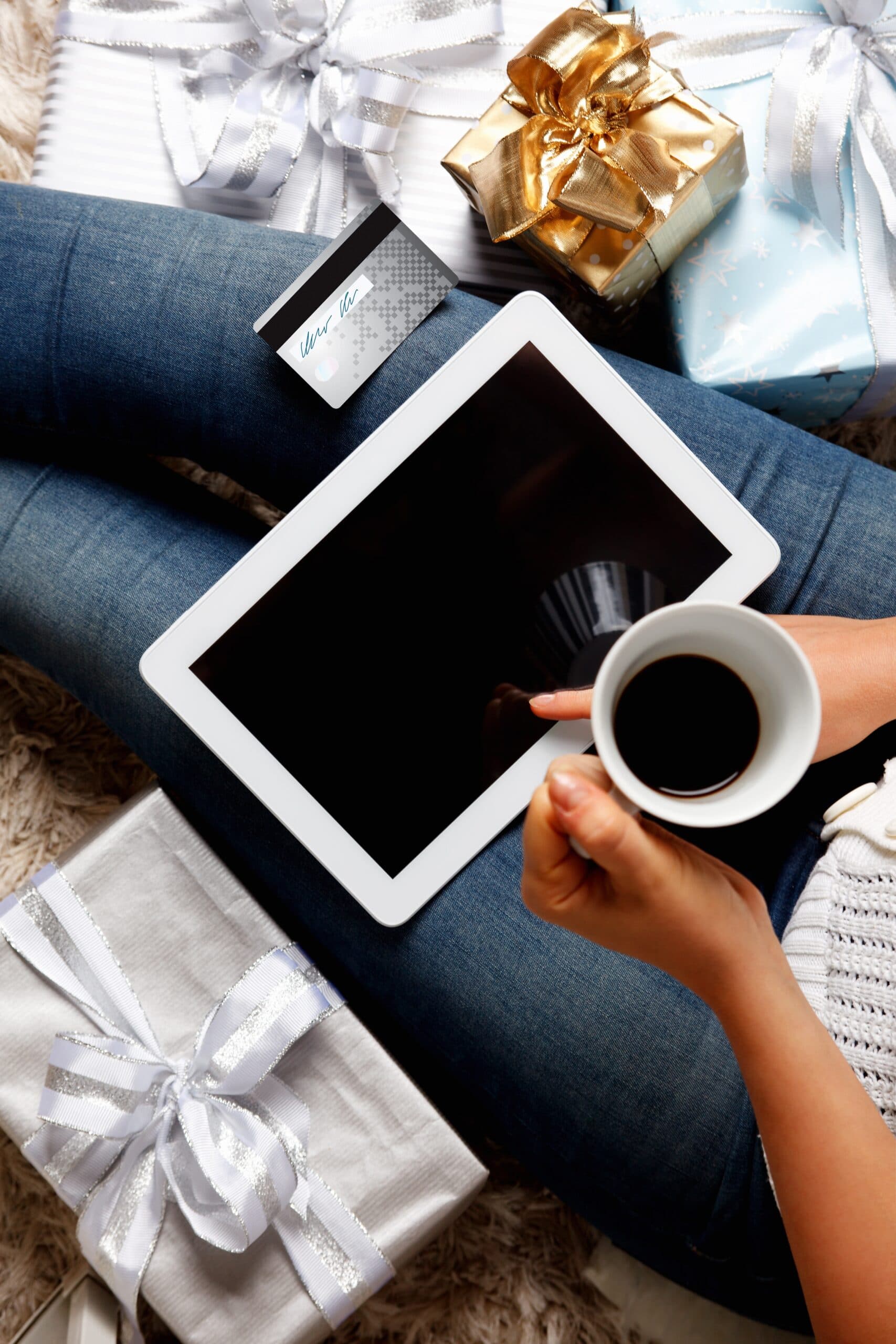 Person sitting with wrapped gifts around, holding a cup of coffee and a tablet on their lap, with a credit card visible, suggesting online shopping or gift planning.