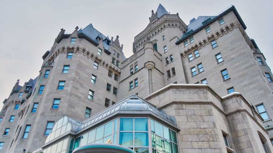 A low-angle view of a large historic stone building with castle-like architecture, featuring turrets, peaked roofs, and numerous windows. Modern glass windows are visible at the ground level.