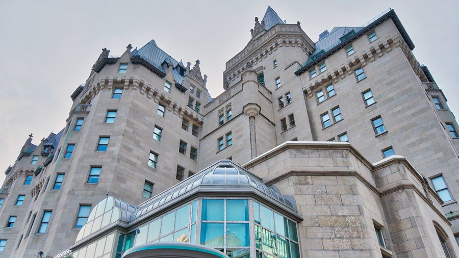 Stone building with castle-like architecture, featuring turrets, pointed roofs, and decorative details, set against a pale sky. Lower part has modern glass windows and a metallic roof accent.