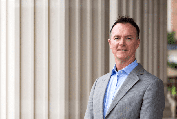 A man in a light gray suit and blue shirt stands outside in front of tall white columns, smiling at the camera.