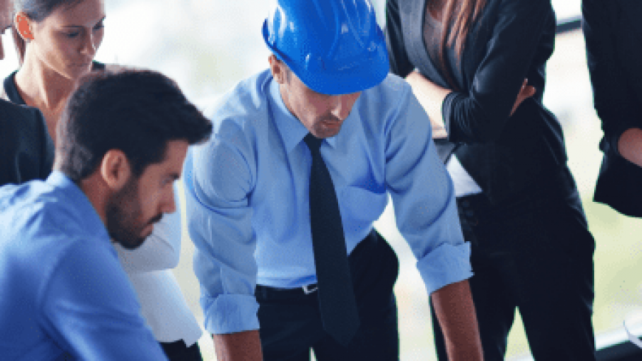 Five professionals, including one person in a blue safety helmet, closely examine blueprints or plans on a table in a bright office, appearing focused and engaged in discussion.
