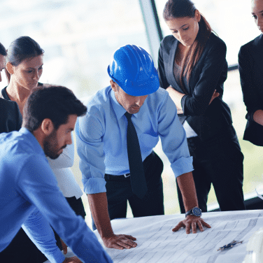 Five professionals, including one person in a blue safety helmet, closely examine blueprints or plans on a table in a bright office, appearing focused and engaged in discussion.