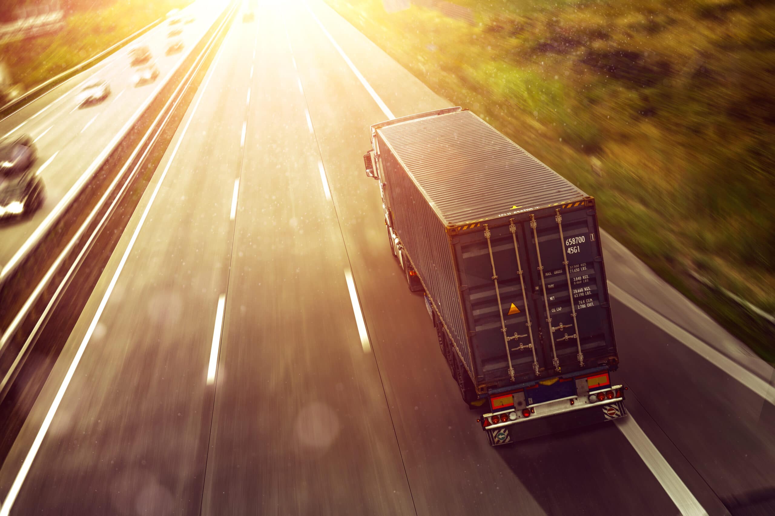 A shipping container truck drives along a sunlit highway, with blurred cars and greenery visible on the left side. The scene is bright, with sunlight illuminating the road and creating reflections on the truck and pavement.