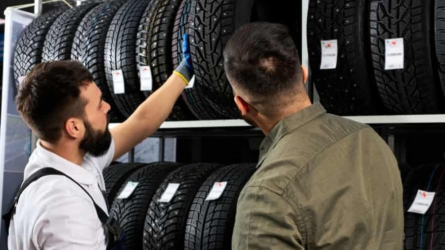 Two men examine and discuss rows of car tires displayed on shelves in a store. One man points at a tire while the other looks on. Each tire has a white price or information tag attached.