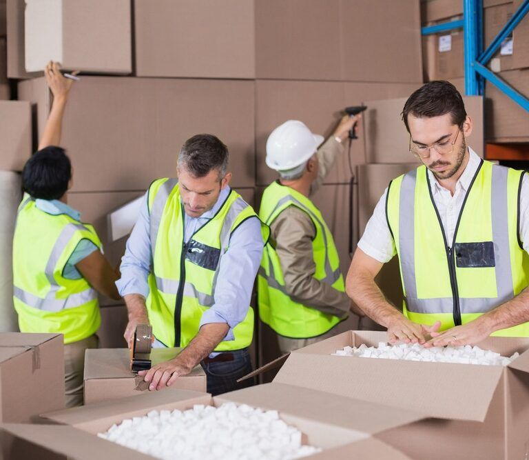 Four workers in safety vests organize and pack boxes in a warehouse. One person tapes a box, another fills a box with packing peanuts, and two others stack boxes on shelves.