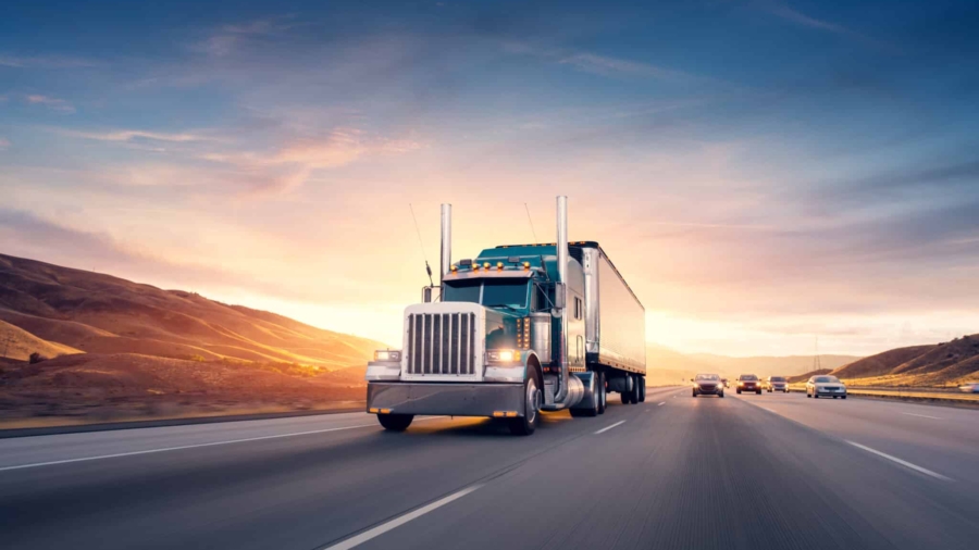 A large semi-truck drives down an open highway at sunset, with hills in the background and several cars traveling in adjacent lanes under a colorful sky.