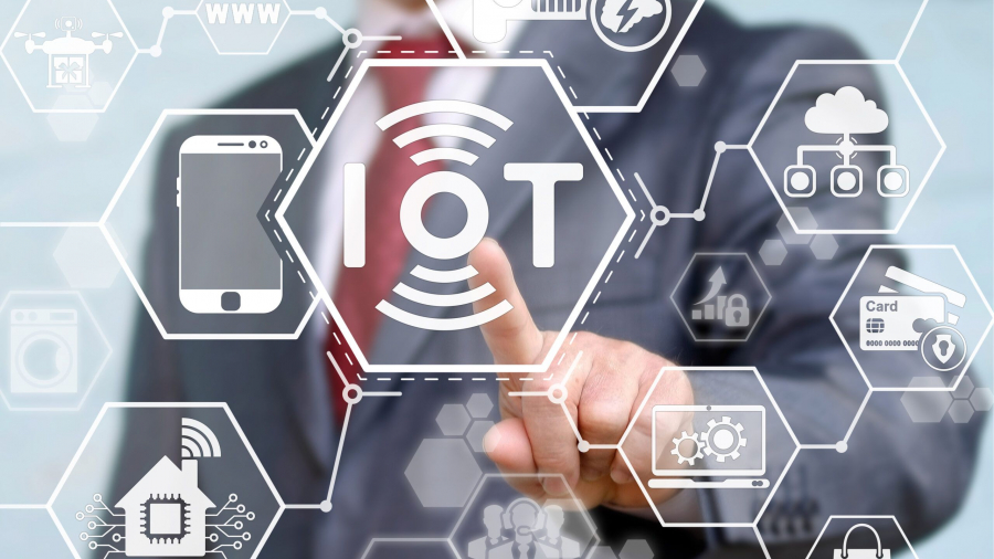 A person in a suit touches a transparent screen with “IoT” at the center, surrounded by icons representing smart devices and technologies, symbolizing the Internet of Things concept.