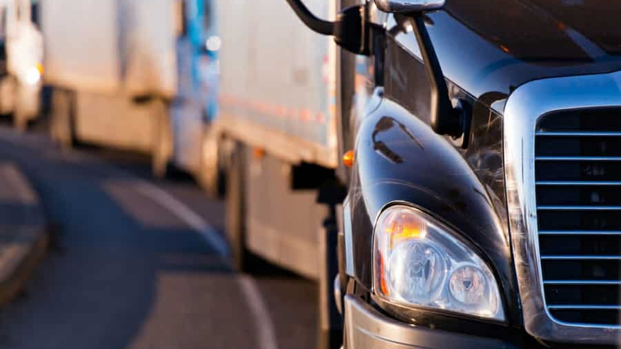 Close-up view of the front of a black semi-truck driving on a curved road, with other trucks visible in the background, all traveling in the same direction.