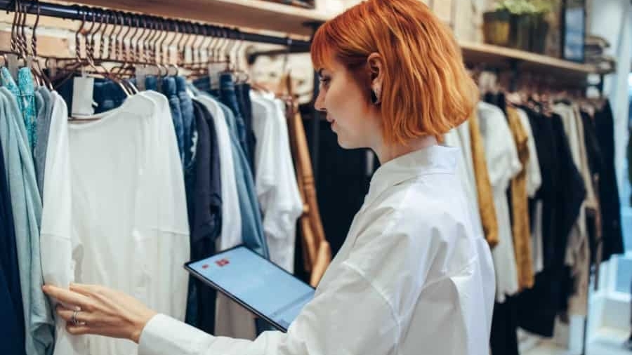 A woman with short red hair in a white shirt holds a tablet while inspecting clothes on a rack in a boutique clothing store.