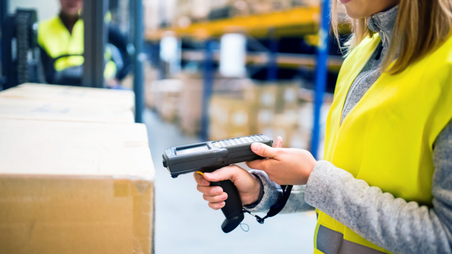 A woman wearing a yellow safety vest uses a handheld barcode scanner to scan boxes in a warehouse. Shelves with more boxes are visible in the background.