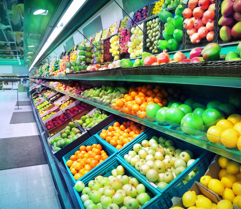 A supermarket produce section with rows of neatly organized fresh fruits and vegetables in baskets and crates, including apples, oranges, lemons, pears, and more, under bright lighting.