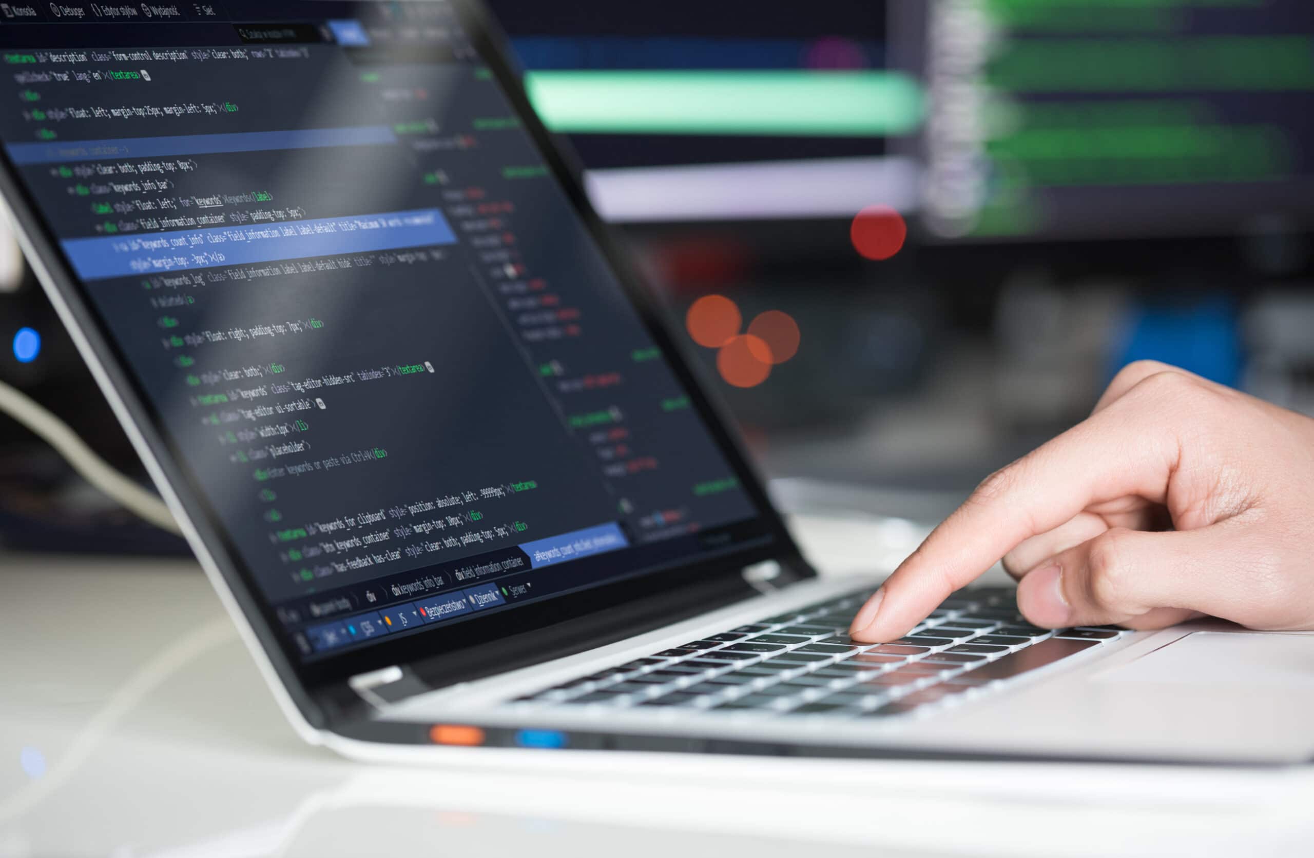 A close-up of a person’s hand typing on a laptop keyboard with programming code displayed on the screen; blurred monitors with code are visible in the background.