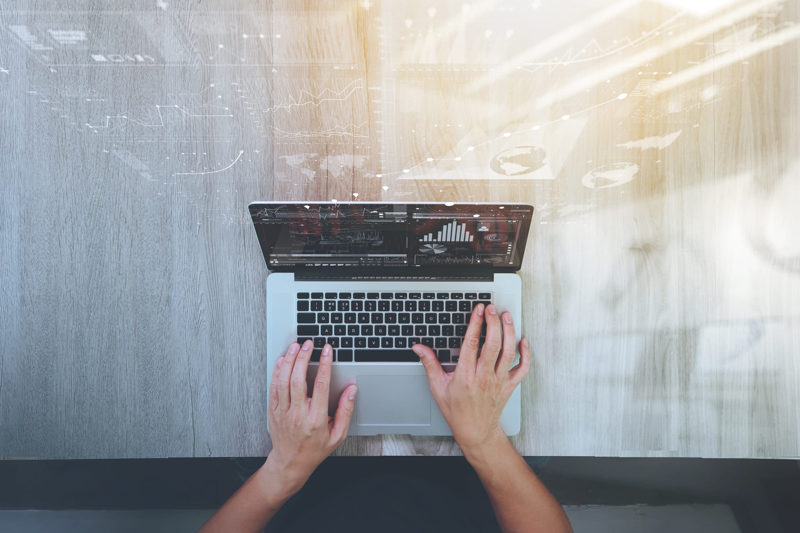 A person types on a laptop at a wooden desk, with data charts and graphs digitally overlaid above the screen, suggesting analysis or business activity. Sunlight shines on the right side of the workspace.