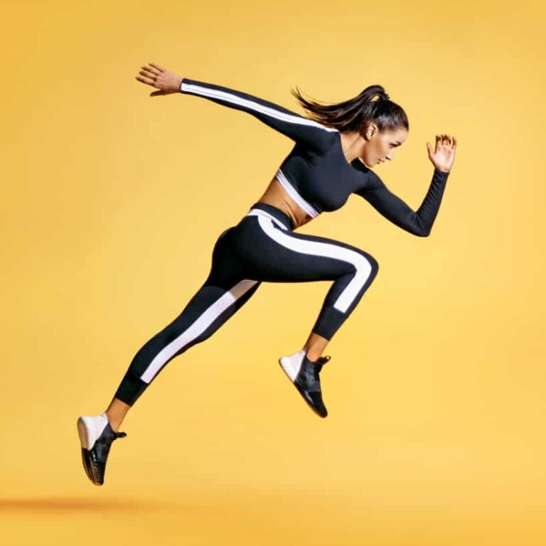 Woman in athletic wear mid-jump against a solid yellow background, appearing energetic and focused, wearing a black and white sports outfit and black sneakers.