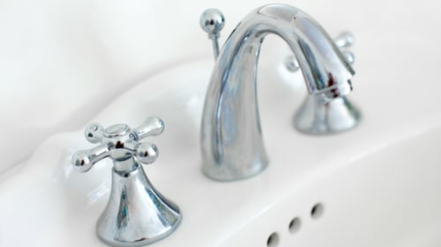 Close-up of a shiny chrome bathroom faucet with two handles on a white porcelain sink. The handles are cross-shaped and the faucet has a curved spout. Three circular overflow holes are visible on the sink.