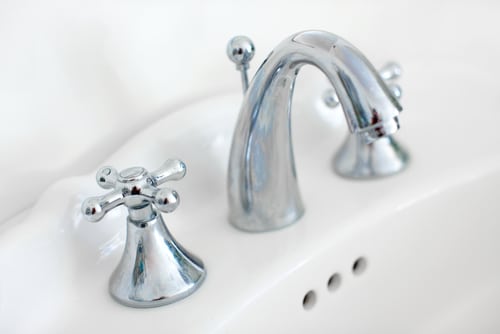 Close-up of a shiny chrome bathroom faucet with two handles on a white porcelain sink. The handles are cross-shaped and the faucet has a curved spout. Three circular overflow holes are visible on the sink.