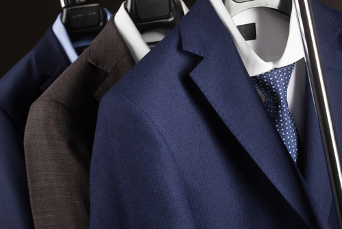 Three suits hanging on hangers, featuring a blue suit with a white shirt and dotted tie in front, followed by a brown and a dark blue suit in the background.