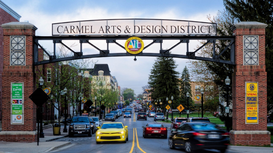 A street view of the Carmel Arts & Design District entrance with a large arched sign overhead, cars driving by, trees, brick pillars, and city buildings visible in the background.