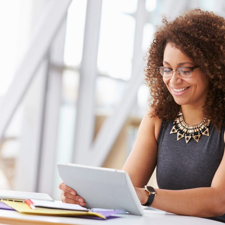 A woman with curly hair and glasses sits at a desk, smiling while using a tablet powered by Power Platform. Papers, notebooks, and a laptop are on the desk. She wears a sleeveless top and statement necklace in a bright, modern setting.