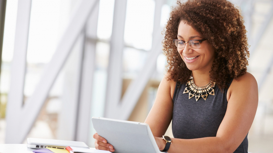 A woman with curly hair and glasses sits at a desk, smiling while using a tablet powered by Power Platform. Papers, notebooks, and a laptop are on the desk. She wears a sleeveless top and statement necklace in a bright, modern setting.