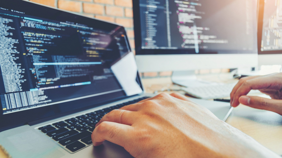 A person types on a laptop with code displayed on the screen. Two large monitors, also showing code, are on a desk in the background—a modern workspace ideal for tech roles like those at enVista Careers.