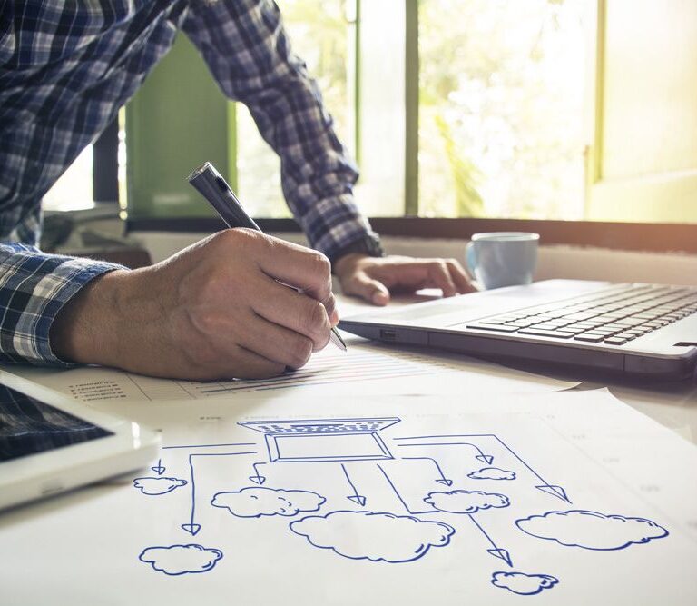 A person writing on paper with cloud diagrams, next to a laptop and tablet running Dynamics 365 Business Central, in a bright office with large windows.