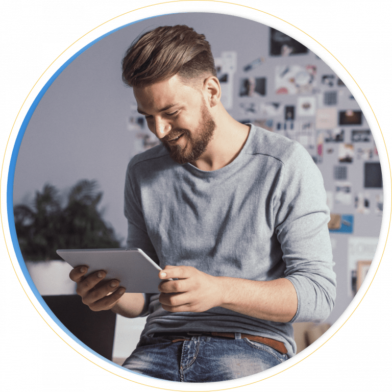 A smiling man with a beard and casual clothes sits indoors, looking at a tablet. A blurred background shows a wall with photos and a plant, as he explores Dynamics 365 Project Operations for managing his projects efficiently.