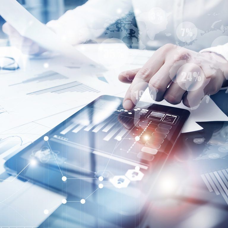 A person reviews financial charts on paper and a digital tablet at a desk, surrounded by graphs and analytics overlays, representing business analysis or data-driven decision-making with advanced technology solutions.