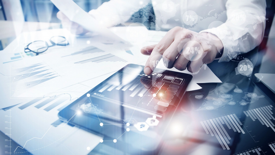 A person reviews financial charts on paper and a digital tablet at a desk, surrounded by graphs and analytics overlays, representing business analysis or data-driven decision-making with advanced technology solutions.