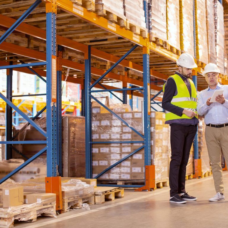 Two men wearing hard hats stand in a warehouse beside tall shelves stacked with boxes. One man in a safety vest and the other with a clipboard discuss inventory, possibly reviewing data from their labor management system.