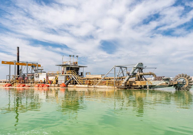 A large industrial dredging machine floats on greenish water under a partly cloudy sky, with pipes, equipment, and a paddle wheel visible on the barge.