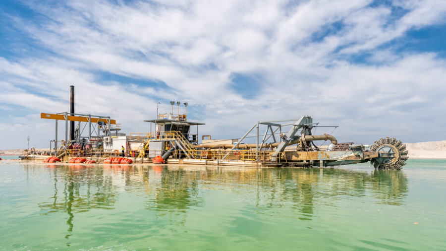 A large industrial dredging machine floats on greenish water under a partly cloudy sky, with pipes, equipment, and a paddle wheel visible on the barge.