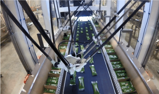 Overhead view of an automated robotic arm in a factory sorting green packaged products on a conveyor belt, surrounded by machinery and industrial equipment.