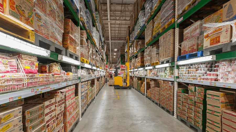 A wide aisle in a warehouse-style grocery store features tall shelves stacked with packaged food products, highlighting the importance of 3PL selection for efficient logistics, with a yellow pallet jack parked at the end of the aisle.