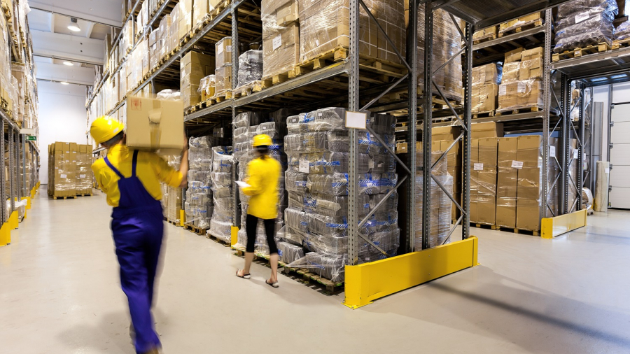 Two workers wearing yellow helmets and uniforms move boxes in a brightly lit warehouse filled with shelves stacked with pallets of goods and cardboard boxes.