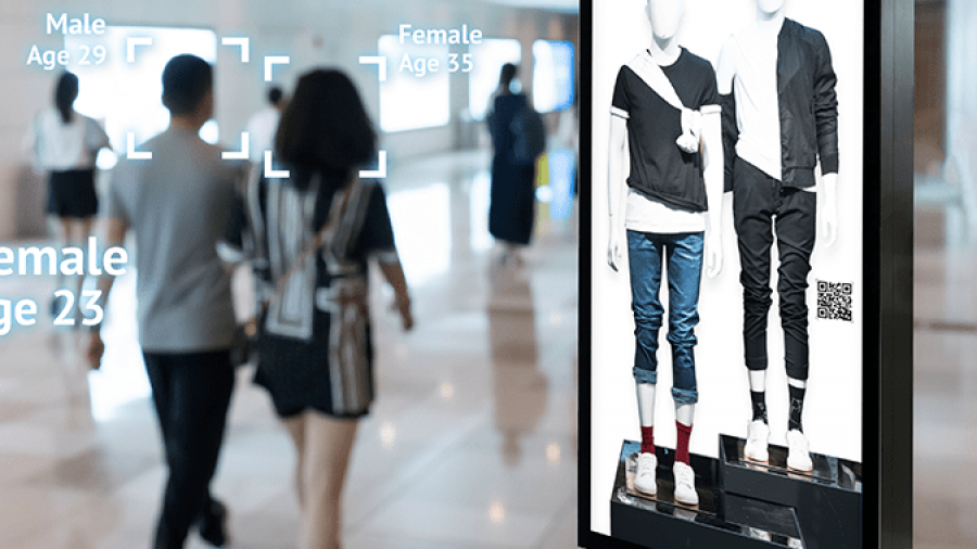 A digital display in a shopping mall shows two mannequins wearing trendy clothing. In the background, facial recognition technology analyzes shoppers, displaying estimated genders and ages above their heads.