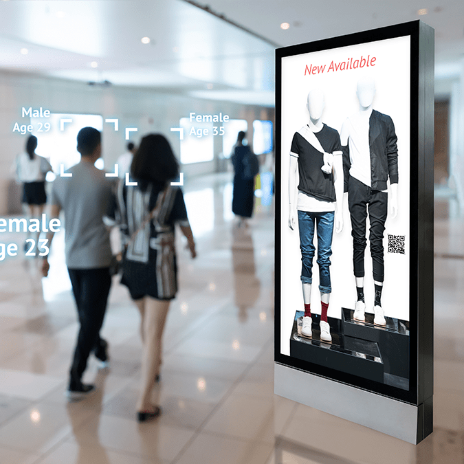 A digital display in a shopping mall shows two mannequins wearing trendy clothing. In the background, facial recognition technology analyzes shoppers, displaying estimated genders and ages above their heads.