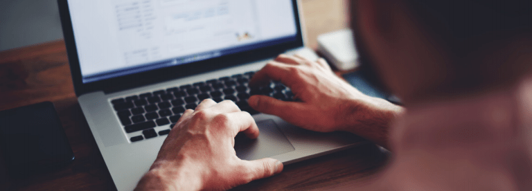 A person uses a laptop at a wooden desk, typing with both hands. The screen displays documents or spreadsheets, and a smartphone lies nearby. The focus is on the hands and laptop keyboard.