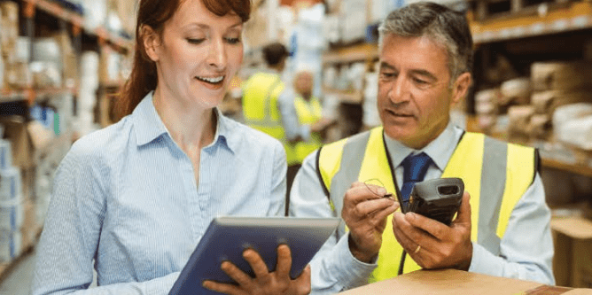 A woman holding a tablet and a man in a safety vest using a handheld device are talking in a warehouse filled with shelves and boxes.