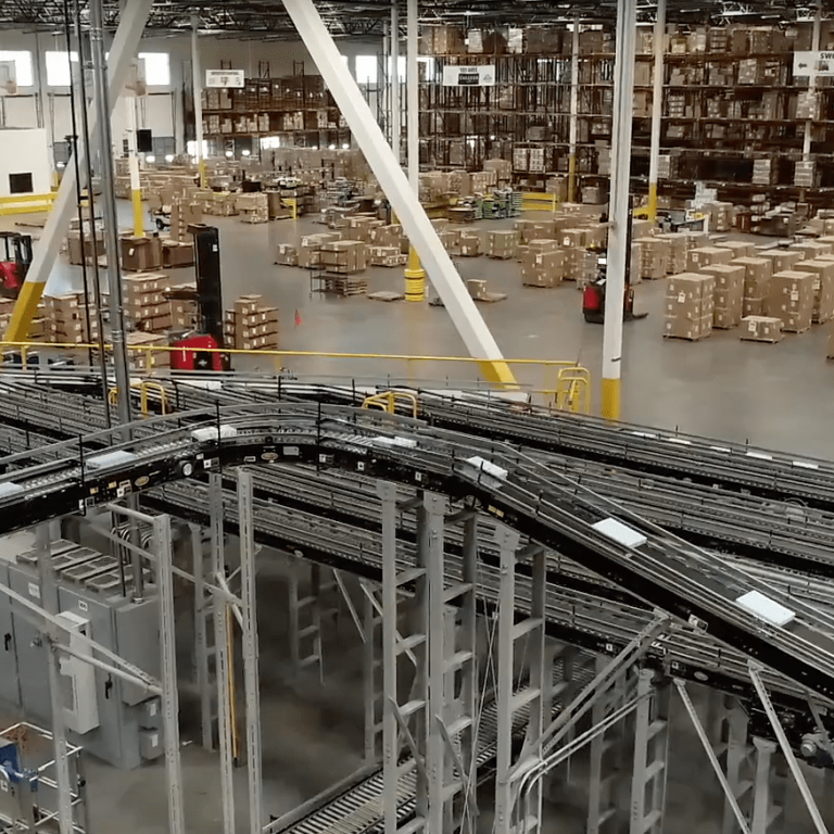 Large warehouse interior with tall shelves stacked with boxes, automated conveyor belts in the foreground, and several forklifts moving pallets around the spacious floor.