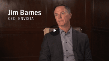 A man in a suit sits for an interview against a dark wood background. Text on the image reads Jim Barnes, CEO, Envista. A play button is centered on the image, indicating a video.
