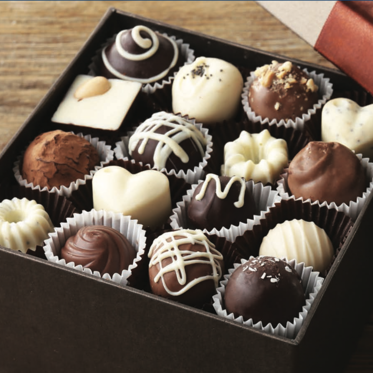 An open box of assorted chocolates in various shapes and colors, including hearts, domes, and squares, with decorative toppings, arranged neatly in paper cups on a wooden surface.