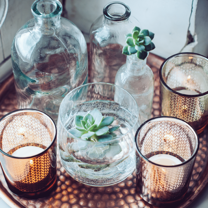 A decorative tray displays glass bottles with succulents inside, a glass bowl with a floating succulent, and three lit candle holders, creating a cozy and elegant arrangement.